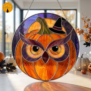 May include: A circular stained glass suncatcher featuring an owl face integrated with a pumpkin design. The piece uses orange, purple, green, and black glass segments, including a small black witch hat detail, suspended by a metal chain.