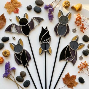 May include: Five stained glass bat ornaments on black metal stakes. The bats feature black, grey, and yellow glass pieces joined with silver solder. They are arranged in various poses, including wings spread, wings folded, and hanging upside down.