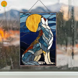 May include: Stained glass panel showing a howling wolf against a yellow moon and a dark blue night sky. The wolf is in shades of white, blue, and brown. The panel is framed and hangs from a chain, suitable for window or wall decor.