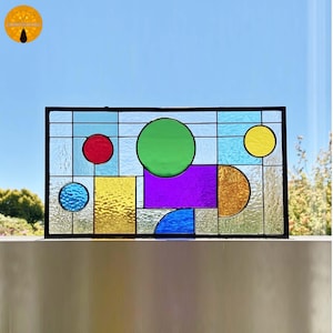 May include: A rectangular stained glass panel with a black border. The design features geometric shapes in various colors, including red, green, yellow, blue, and purple. The panel is displayed against a bright blue sky.