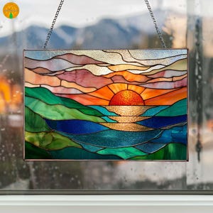 Puede incluir: Panel de vidrieras que representa una puesta de sol sobre el océano. La obra de arte presenta una vibrante paleta de colores naranja, rojo, verde y azul. El sol es un orbe naranja brillante y el océano es una mezcla de azul y dorado. El panel está enmarcado y cuelga de una cadena.