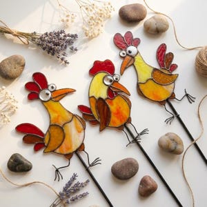 Könnte beinhalten: Drei Gartenstecker aus Buntglas in Form von Hühnern, bestehend aus gelben, orangen und roten Glassegmenten mit dunklen Metalllötstellen. Jedes Huhn hat einen roten Kamm, einen spitzen Schnabel und runde Augen, befestigt an einem schwarzen Metallstab.