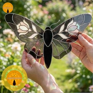 May include: Stained glass butterfly in shades of black, grey, and white. The wings have floral patterns and are held up by hands. The butterfly has a black body and is made of multiple glass pieces.