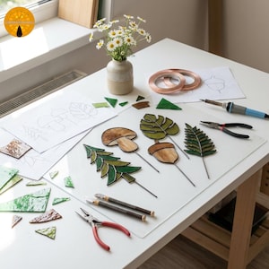 Puede incluir: Una mesa blanca exhibe materiales para el arte del vitral. La escena incluye trozos de vidrio, alambre de cobre, alicates, un soldador y dibujos de hojas, setas y bellotas. Un pequeño jarrón contiene margaritas.