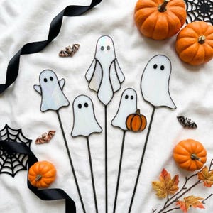 May include: Five stained glass ghost ornaments on thin metal stakes, featuring white iridescent glass with black solder lines for eyes. One ghost holds a small orange glass pumpkin. The set includes three small orange pumpkins and two metal bat charms.