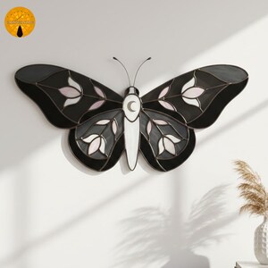 May include: A stained glass butterfly wall hanging. The butterfly features black, gray, white, and pink glass pieces. The wings have a floral design. The body is white with a crescent moon. The antennae are thin and black.