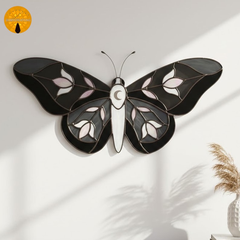 May include: A stained glass butterfly wall hanging. The butterfly features black, gray, white, and pink glass pieces. The wings have a floral design. The body is white with a crescent moon. The antennae are thin and black.
