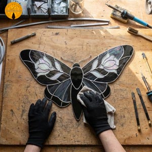 May include: A stained glass butterfly with black and white wings and a black body. The butterfly is being cleaned with a cloth. The wings feature a floral design. Tools and materials for stained glass art are in the background.