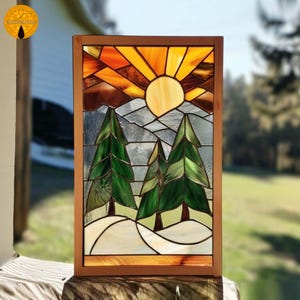 May include: Stained glass panel featuring a mountain scene with a setting sun and evergreen trees. The artwork has a wooden frame and showcases a vibrant color palette of orange, yellow, green, and gray. The design evokes a sense of nature and tranquility.