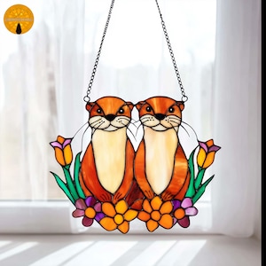 Pode incluir: Arte em vitral com duas lontras com pelagem laranja e barrigas brancas. As lontras estão rodeadas por flores e folhagens coloridas, com uma corrente para pendurar. A obra de arte foi concebida para ser exibida numa janela.