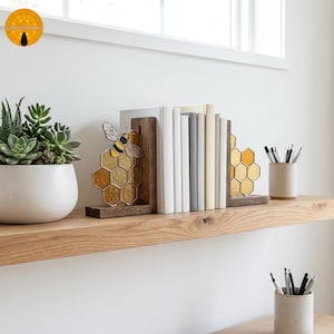 Puede incluir: Estante de madera contra una pared blanca. Muestra una maceta de cerámica blanca con una planta suculenta, libros sujetados por sujetalibros con temática de abeja y una pequeña taza de cerámica con bolígrafos y lápices.