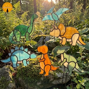 May include: A collection of six stained glass dinosaur garden stakes featuring a green long-necked dinosaur, a blue stegosaurus, an orange tyrannosaurus, a yellow triceratops, a light green parasaurolophus, and a light blue flying pterodactyl.