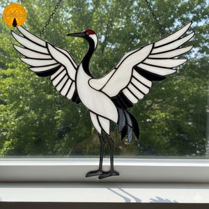 May include: Stained glass crane with outstretched wings, featuring white, black, and red accents. The bird's body is primarily white, with black wingtips and tail feathers. The crane is suspended by a chain, creating a decorative window hanging.
