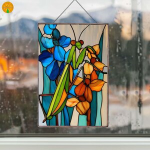 May include: Stained glass panel featuring a praying mantis amidst blue and orange flowers. The artwork has a rectangular shape, framed with a metal border, and is suspended by a chain. The flowers and insect are rendered in vibrant colors, creating a decorative piece.