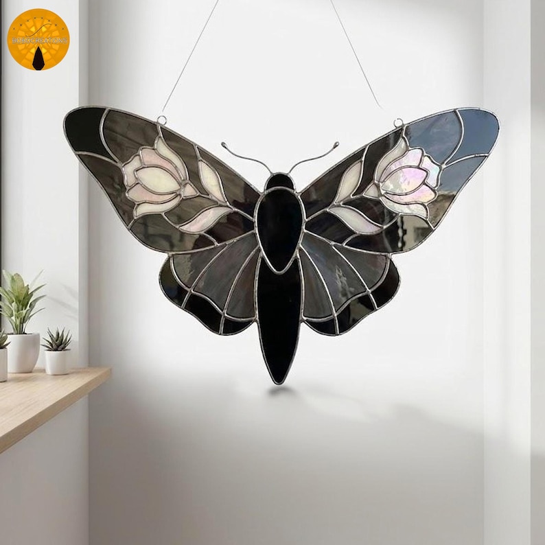 May include: A stained glass butterfly with a black body and wings in shades of gray, white, and iridescent pink. The wings feature floral designs. The artwork is suspended by a thin wire, suitable for wall decor.