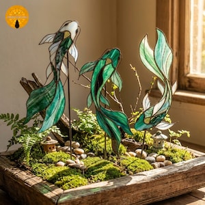 May include: Three stained glass koi carp sculptures in green and white are displayed in a wooden planter filled with moss, small stones, and miniature Japanese lanterns. The fish are mounted on metal rods.