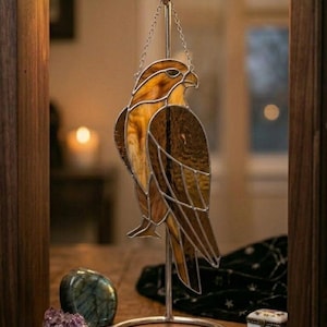 May include: A stained glass hawk sculpture in shades of brown and amber, suspended from a silver chain. The bird is mounted on a silver stand. The artwork is displayed on a wooden shelf with decorative stones and a small trinket box.