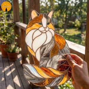 Peut inclure: Un attrape-soleil en vitrail en forme de chat assis avec une longue queue recourbée. Le chat présente une mosaïque de pièces de verre blanc, ambre, orange et gris assemblées par des lignes de soudure en métal sombre, formant une silhouette féline.
