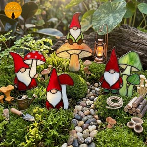 Op de afbeelding: Een verzameling glas-in-lood tuinkabouters en paddenstoelen in een weelderige, groene omgeving. De kabouters hebben rode hoeden en witte baarden, sommige zitten op kleurrijke paddenstoelenkappen. Een kleine lantaarn en miniatuur tuingereedschap dragen bij aan de grillige scène.