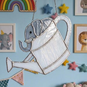 May include: A stained glass elephant in a watering can design, hanging from a chain. The elephant is gray and white, with a black eye. The watering can is white with silver accents. The background is a light blue wall with decorative items.