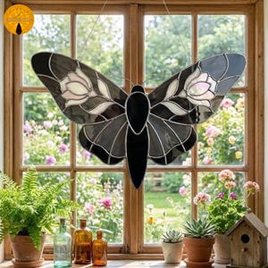 May include: A stained glass butterfly suncatcher with black, gray, and white details. The wings feature floral designs. The suncatcher hangs in front of a window with a garden view, adding a decorative touch to any space.