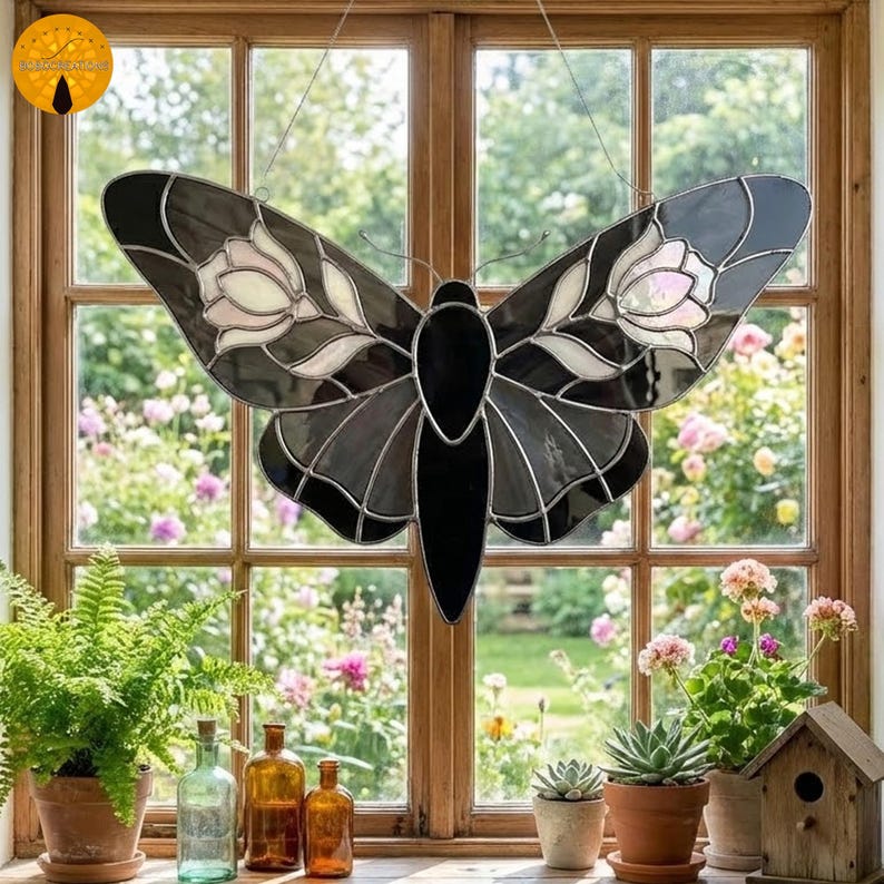 May include: A stained glass butterfly suncatcher with black, gray, and white details. The wings feature floral designs. The suncatcher hangs in front of a window with a garden view, adding a decorative touch to any space.