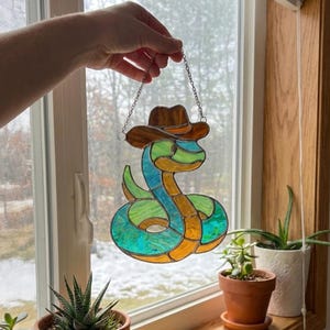 Può includere: Vetrata a forma di serpente con cappello da cowboy marrone, con segmenti blu, verdi e arancioni. Il serpente è sospeso da una catena d'argento. La finestra mostra alberi e un cielo nuvoloso.