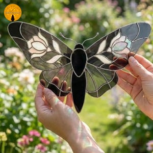 May include: A stained glass butterfly with black, gray, and white details. The wings feature floral designs and are held up against a blurred background of green foliage and flowers. The butterfly is a decorative item.