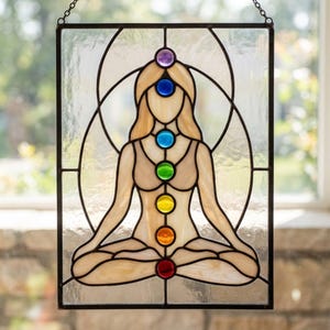 May include: Stained glass panel featuring a meditating figure with seven colored chakra symbols. The figure is in a seated lotus position, with a black metal frame and chain for hanging. The glass has a beige and clear color scheme.