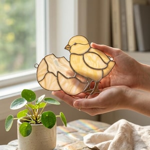 May include: A stained glass chick emerging from its egg. The chick is yellow with a detailed beak and feet. The eggshell is a creamy white color. The piece is held in front of a window with a small potted plant in the foreground.
