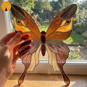 Puede incluir: Atrapasoles de mariposa de vidrieras con tonos marrones, naranjas y amarillos. Las alas presentan patrones detallados. La mariposa está diseñada con un lazo de metal para colgar. Un artículo decorativo.