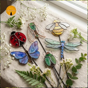 Pode incluir: Uma coleção de estacas de jardim de vitrais coloridos em forma de insetos, incluindo uma joaninha, uma borboleta, uma abelha, uma libélula, uma cigarra e um besouro. As estacas estão dispostas sobre uma superfície de linho neutra com flores brancas e vegetação.