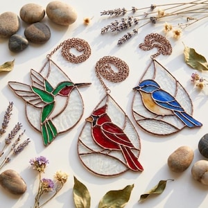 Peut inclure: Trois capteurs de soleil en vitrail représentant un colibri vert, un cardinal rouge et un oiseau bleu, chacun enfermé dans un cadre en forme de goutte avec une bordure en métal cuivré et une chaîne de suspension assortie fixée au sommet.