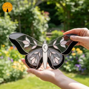 May include: A stained glass butterfly with a black, gray, white, and pink color scheme. The butterfly has a crescent moon detail and floral patterns on its wings. The artwork is held in front of a blurred green background.