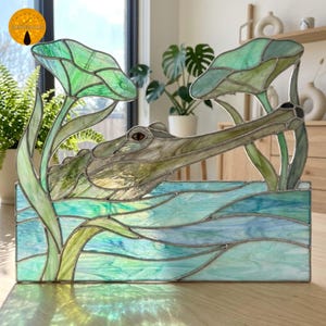 May include: A stained glass panel featuring a gharial crocodile with a long snout emerging from blue and green water, surrounded by tall green lily pads. The piece is constructed with soldered metal lines and various shades of textured glass.