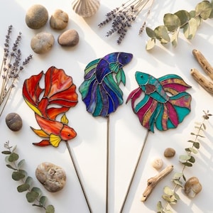 May include: Three stained glass betta fish ornaments on metal stakes. One fish is red and yellow, one is blue and purple, and one is teal and magenta. Each fish features intricate glass segments held together by dark metal solder lines.