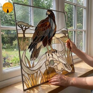 May include: A rectangular stained glass panel featuring a brown and black eagle perched atop a white animal skull. The background depicts a savanna landscape with amber-colored grass and trees, all framed in a thin metal border with clear glass.