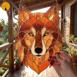 May include: Stained glass wolf head with amber, orange, and cream tones. The artwork features a detailed mosaic design with a black nose and yellow eyes. The piece is designed to hang and is suitable for home decor.