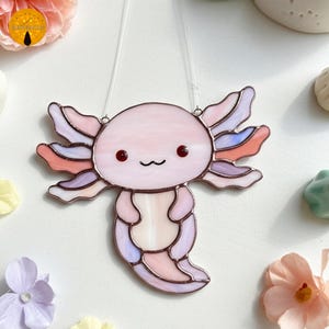 Peut inclure: Un attrape-soleil axolotl en vitrail avec un corps rose, des yeux rouges et des branchies multicolores. L'axolotl a une bouche souriante et est suspendu à une ficelle transparente. Objet décoratif.