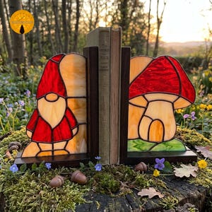 May include: A pair of stained glass bookends featuring a gnome on the left and a mushroom on the right. Each piece is crafted with red, cream, and green glass panels held by dark metal frames, mounted on dark wooden bases to support a book.