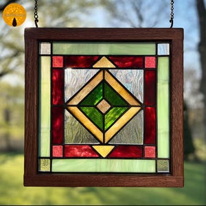 Puede incluir: Panel de vidriera cuadrado con marco de madera marrón oscuro. El diseño presenta un rombo central en verde y dorado, rodeado de vidrio rojo, amarillo y verde claro. El panel está colgado de una cadena.