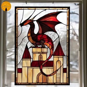 Puede incluir: Panel de ventana de vidrieras con un dragón rojo y naranja posado sobre un castillo con torretas rojas. El dragón tiene grandes alas y está sobre un fondo claro y texturizado. La ventana está enmarcada en negro.