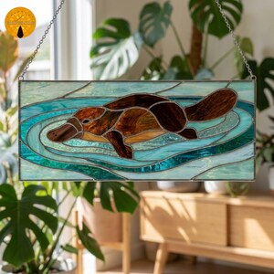 Puede incluir: Un panel rectangular de vidriera que muestra un ornitorrinco marrón nadando en el agua. El diseño utiliza varios tonos de vidrio azul y verde azulado para representar ondas y olas, con vidrio marrón oscuro y bronceado formando el cuerpo.
