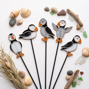 Puede incluir: Cinco adornos de vitral con forma de frailecillo sobre finas varillas de metal negro. Cada frailecillo presenta piezas de vidrio blanco, negro y naranja con líneas de soldadura. Las aves están dispuestas en abanico rodeadas de conchas marinas.