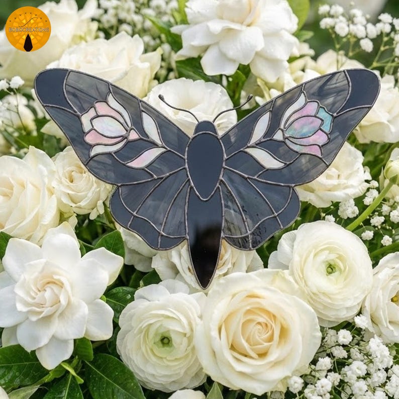 May include: A decorative butterfly with a stained glass design. The butterfly features a dark gray body and wings with pink, white, and light blue accents. The butterfly is set against a backdrop of white roses and greenery.