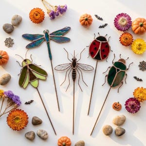 Op de afbeelding: Een verzameling van vijf tuinstekers van glas-in-lood in de vorm van insecten, waaronder een blauwe libel, een groene mot, een mug, een rood lieveheersbeestje en een groene kever, verspreid tussen droogbloemen, pompoenen en vleermuizen.