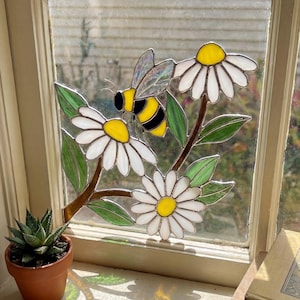 May include: Stained glass window art depicting a bee and daisies. The bee is yellow and black, and the daisies have white petals and yellow centres. The artwork is set in a window frame.