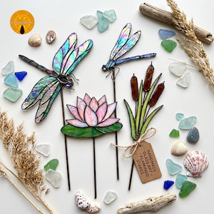 May include: Stained glass garden stakes featuring two dragonflies with iridescent wings, a pink lotus flower on a green lily pad, and a cluster of brown cattails. A brown paper tag attached to the cattails reads HANDMADE GLASS ART - FLAMINGO STAKES.