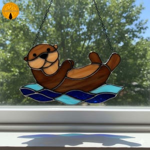 Può includere: Suncatcher in vetro colorato a forma di lontra. La lontra è marrone e beige, che galleggia su onde blu e turchesi. Il suncatcher è appeso a una catena nera. La lontra è in posa rilassata.