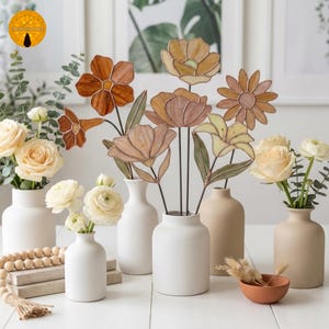 May include: A collection of decorative glass flowers in various shapes and warm colors, including orange, brown, and yellow, displayed in white and beige ceramic vases. Fresh roses and other flowers are also arranged in vases, creating a floral display.